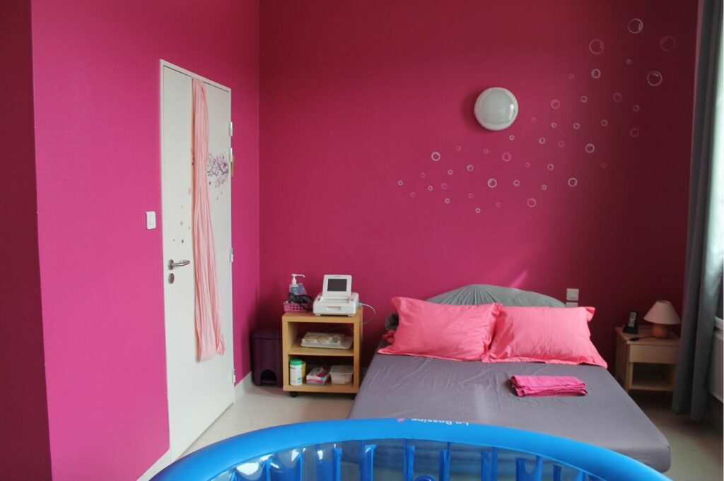 Les chambres de la maison de naissance de Bourgoin-Jallieu sont colorées et accueillantes. Sur la photo, la chambre baigne dans une ambiance rose fuschia et une piscine d’accouchement est présente près du lit double.