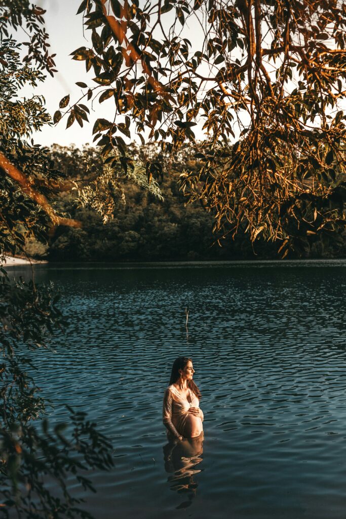 Une femme enceinte regarde le soleil alors qu’elle se baigne au milieu d’un lac, vêtue d’une robe à manches longues. Son ventre arrondi repose sur l’eau calme et bordée d’arbres.